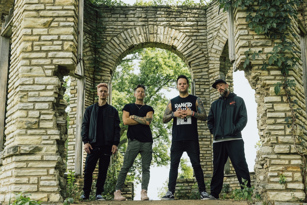 Members of rock band Story of the Year stand outdoors together framing themselves inside stone arch ruins covered in green ivy.